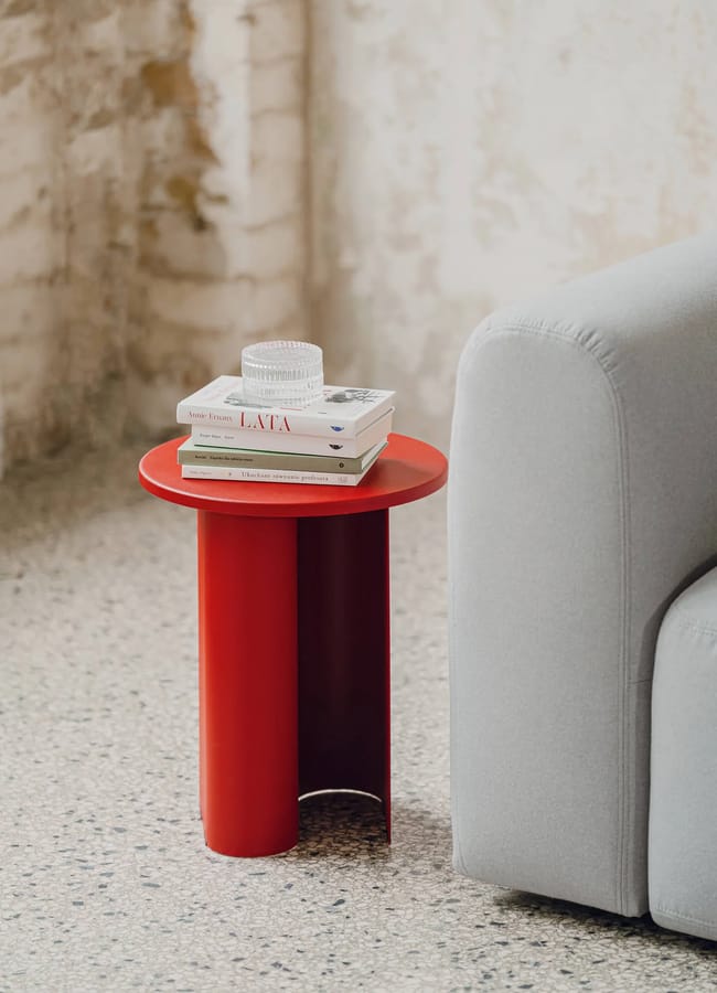 A small red side table, inspired by the Unexpected Red Theory in interior design, sits beside a light grey sofa, holding books and a glass candle holder on a speckled floor against a textured wall.