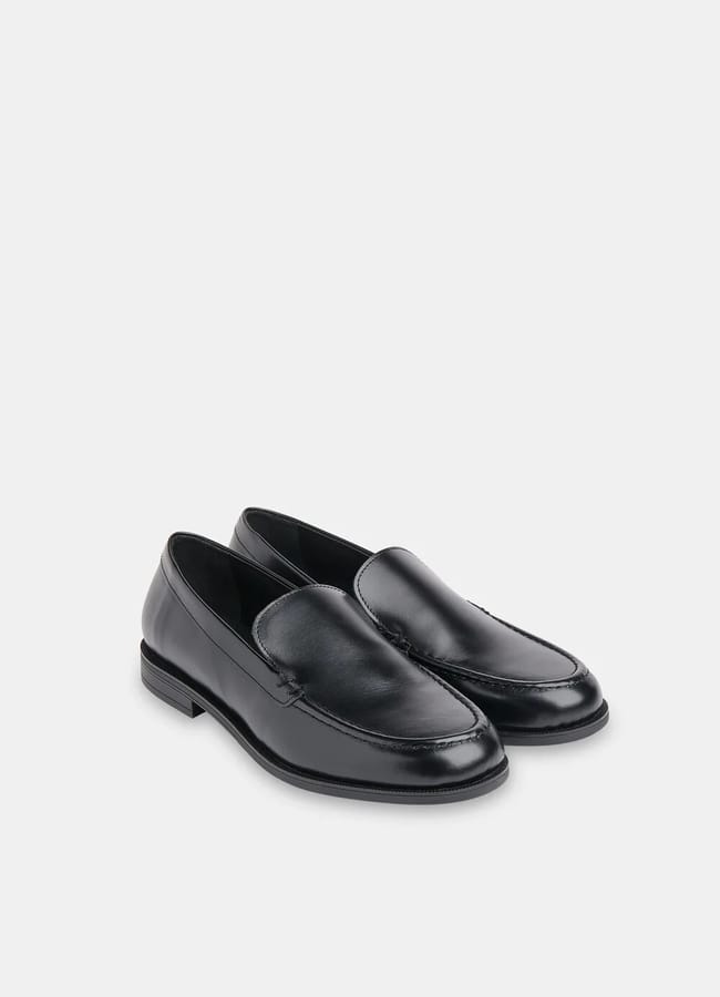 A pair of classic black leather loafers with low heels and rounded toes, perfect for chic fashion when paired with brown trousers, placed on a plain light grey background.