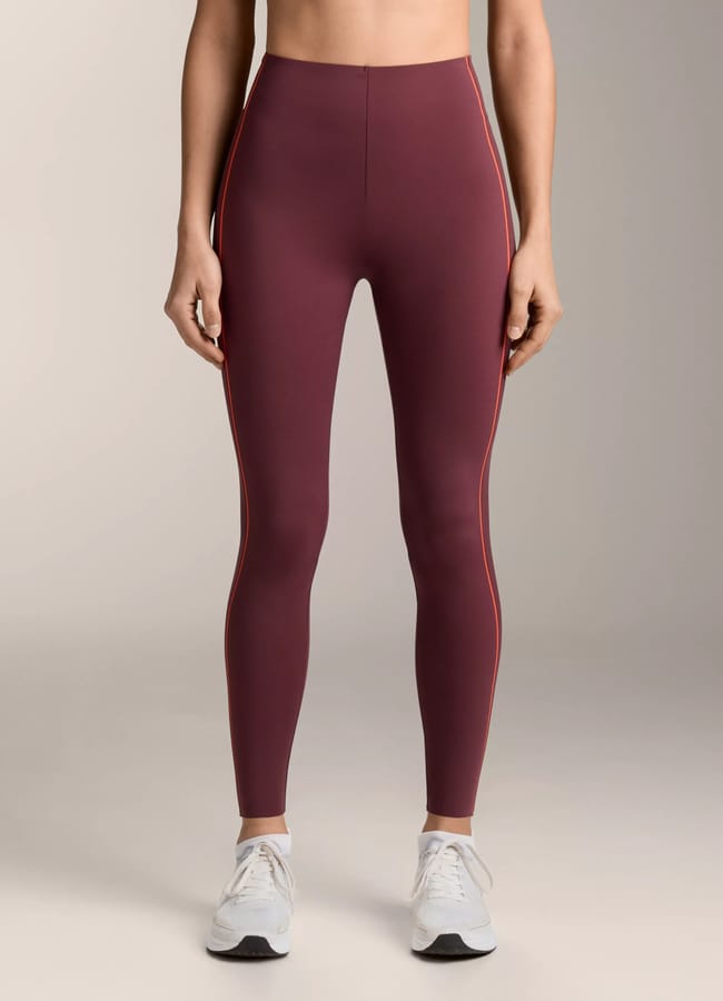 A person stands facing forwards, wearing high-waisted, burgundy leggings with subtle side stripes from the latest activewear collection and white trainers, against a plain, light background. Their face is not visible.