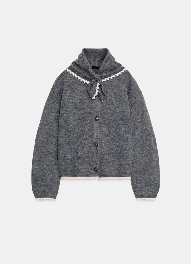 Grey button-up cardigan with long sleeves, white lace trim along the collar, cuffs, and hem, plus a scarf-style collar detail tied at the front. Pair with brown trousers for chic outfits that reflect current fashion trends. Cardigan is laid flat on a plain white background.