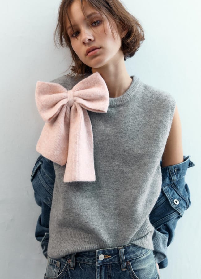 A young person with short brown hair wears a sleeveless grey jumper featuring bow fashion—a large pink bow on the shoulder—layered over a denim jacket and jeans, standing against a plain light background.