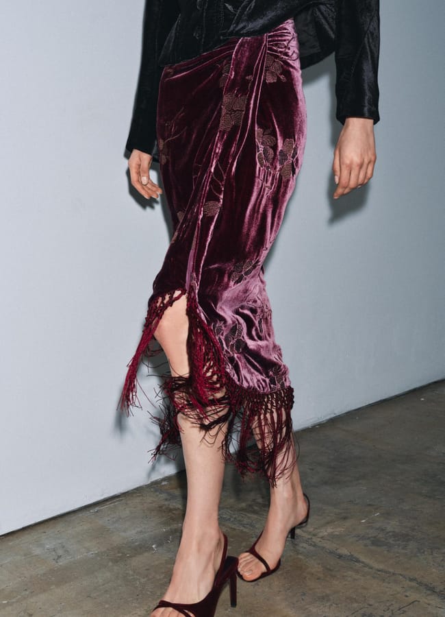 A person wearing a deep burgundy velvet party wear skirt with fringe and lace details, paired with a black long-sleeve top and burgundy high-heeled sandals, stands on a concrete floor against a light grey wall.