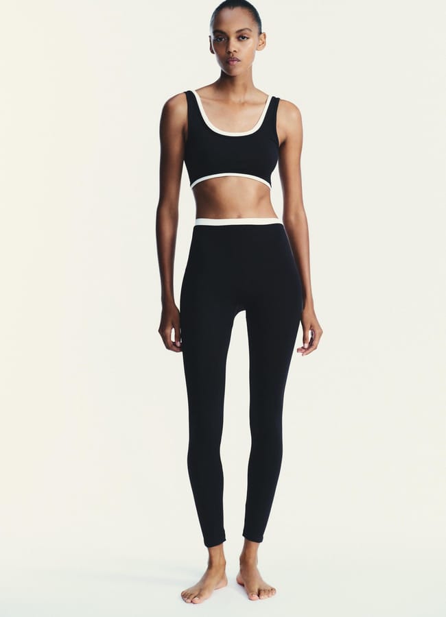 A person stands barefoot against a plain background, wearing the latest activewear: a matching black athletic crop top and high-waisted leggings with white trim along the edges.