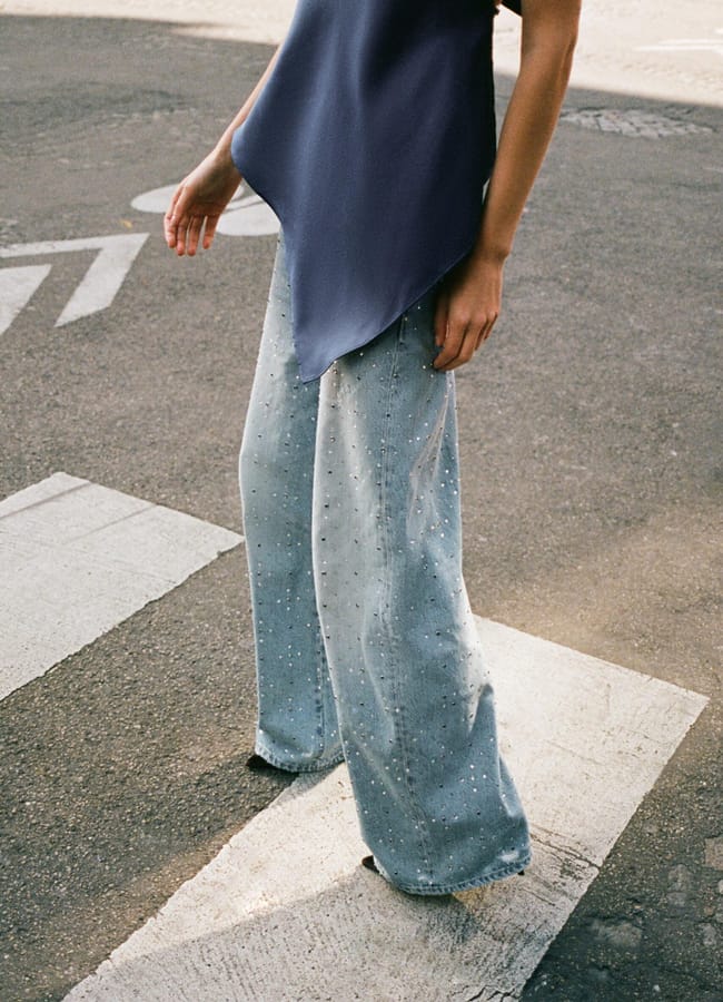 A person stands on a pedestrian crossing in a sleeveless, asymmetrical navy top and light blue embellished jeans with small bead details. Their head is out of frame.