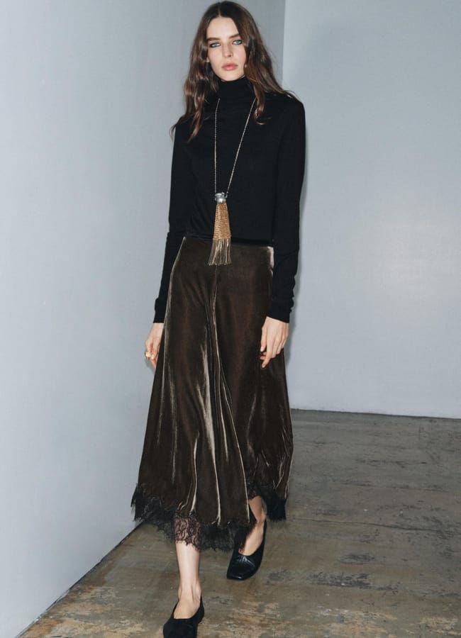 A woman with long brown hair wears a black polo-neck jumper, a long pendant necklace, and a dark brown velvet party-wear midi skirt with lace trim and black shoes, walking indoors on a concrete floor.