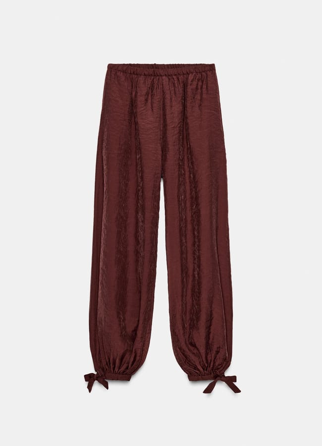 Loose-fitting, shiny maroon trousers inspired by casual brown trousers, featuring an elasticated waistband and gathered ankles tied with small bows, displayed against a plain white background.