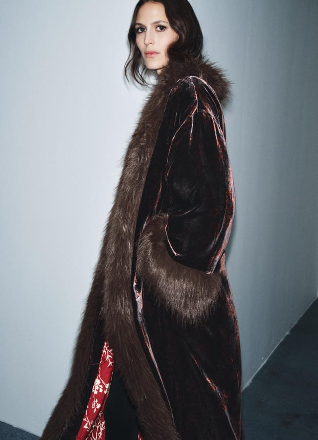 A person with long, wavy dark hair stands against a grey wall, wearing velvet partywear—a dark velvet coat with brown faux fur trim and a glimpse of red patterned lining visible at the bottom.
