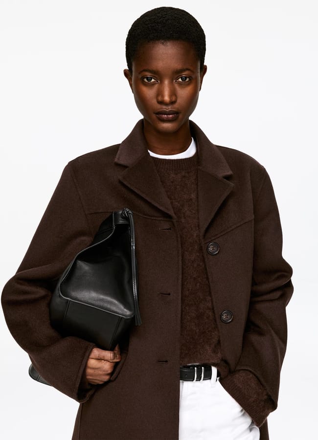 A person stands against a plain background, styled like a Fashion Editor in a dark brown coat over a jumper, white trousers, and holding a black leather bag under their arm.
