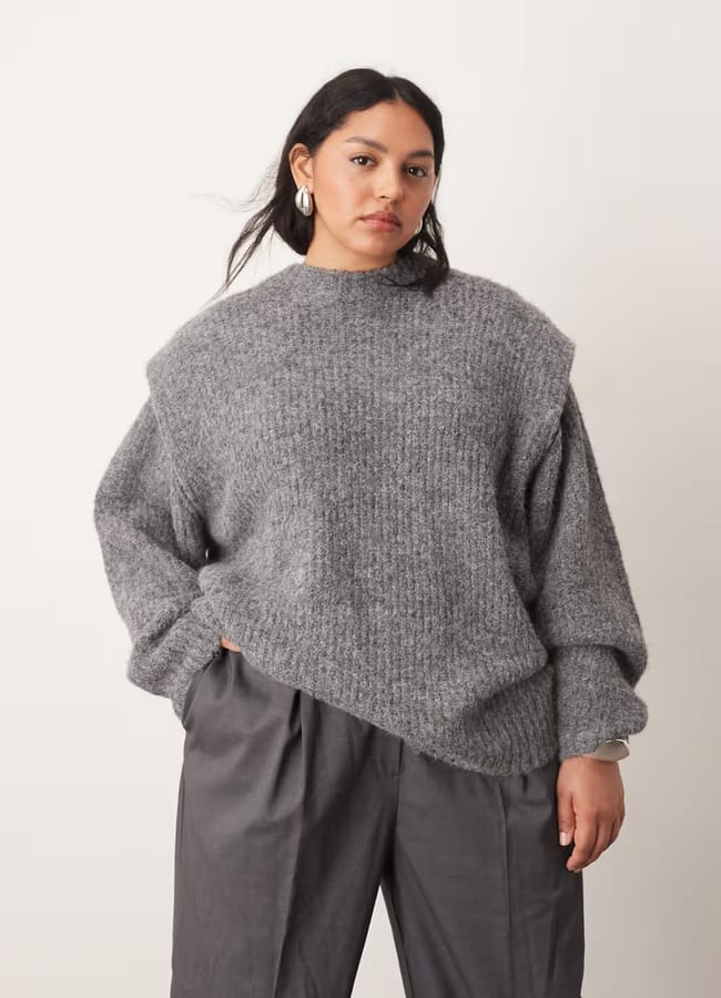 A woman wearing a loose grey knit jumper and wide-leg grey trousers stands against a plain white background, showcasing effortless women's workwear as she looks confidently at the camera.