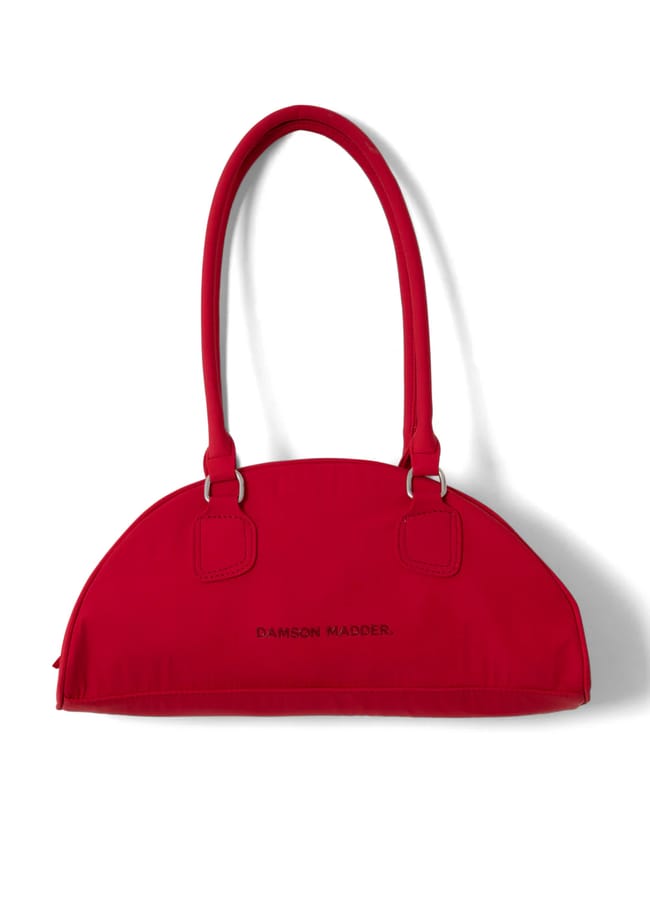 A bright red, semi-circular handbag with two handles and the words DAMSON MADDER printed in black on the front—perfect for those who love bold red accessories. The bag is photographed against a white background.