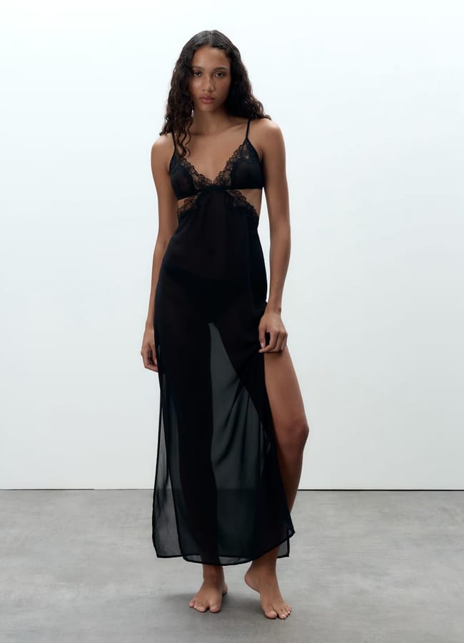 A woman stands barefoot on a grey floor, wearing a long, black, sheer lace lingerie set with spaghetti straps and a thigh-high slit, against a plain white background.