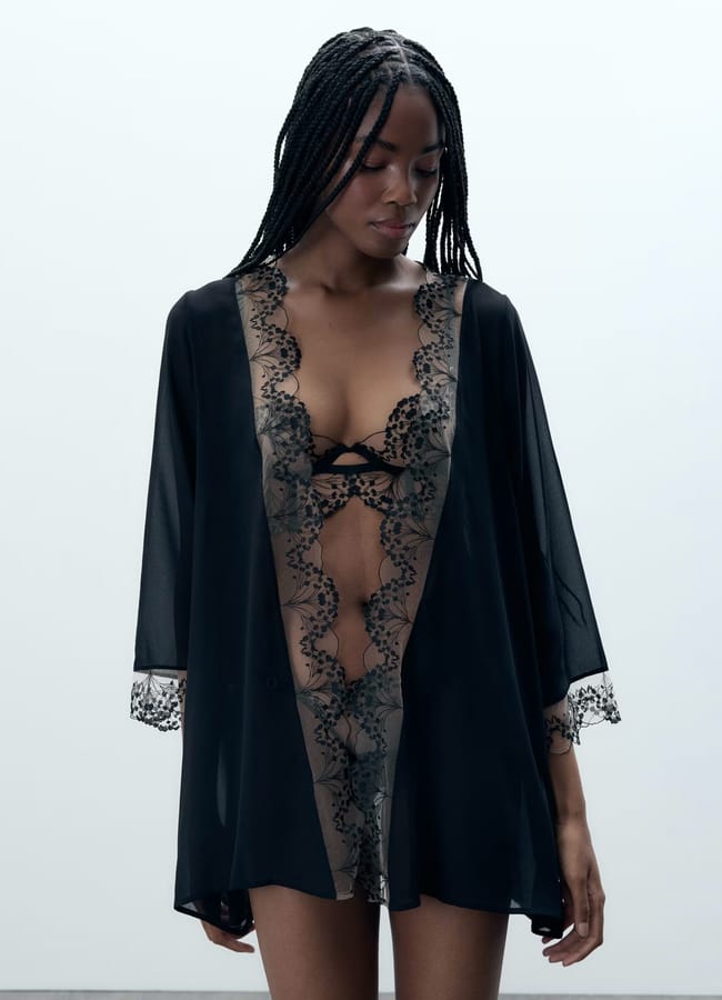 A woman with long plaited hair wears a sheer black lace lingerie set, featuring intricate detailing along the neckline and cuffs, standing against a plain light background and looking down.