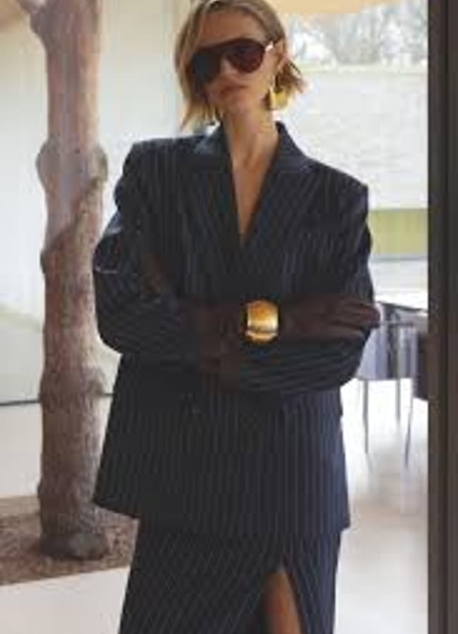 A person, reminiscent of a Fashion Editor, wearing a dark pinstripe suit and gold bracelet stands confidently with arms crossed near a window—embodying the bold style of High Street Blazers.