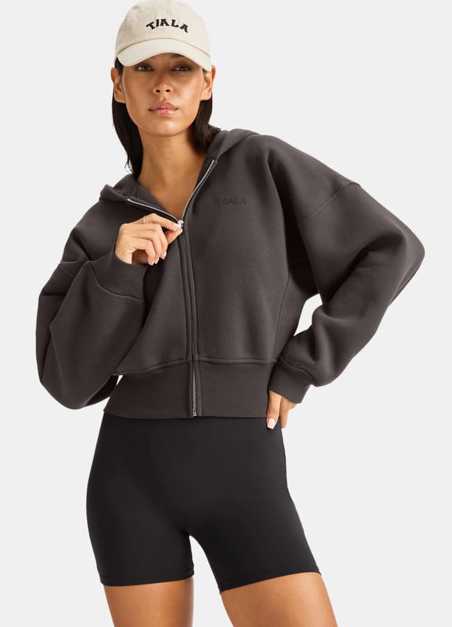 A woman wearing a beige cap, dark grey zip-up athleisure hoodie, and black cycling shorts stands with one hand on her hip and the other holding the zip, looking confidently at the camera.