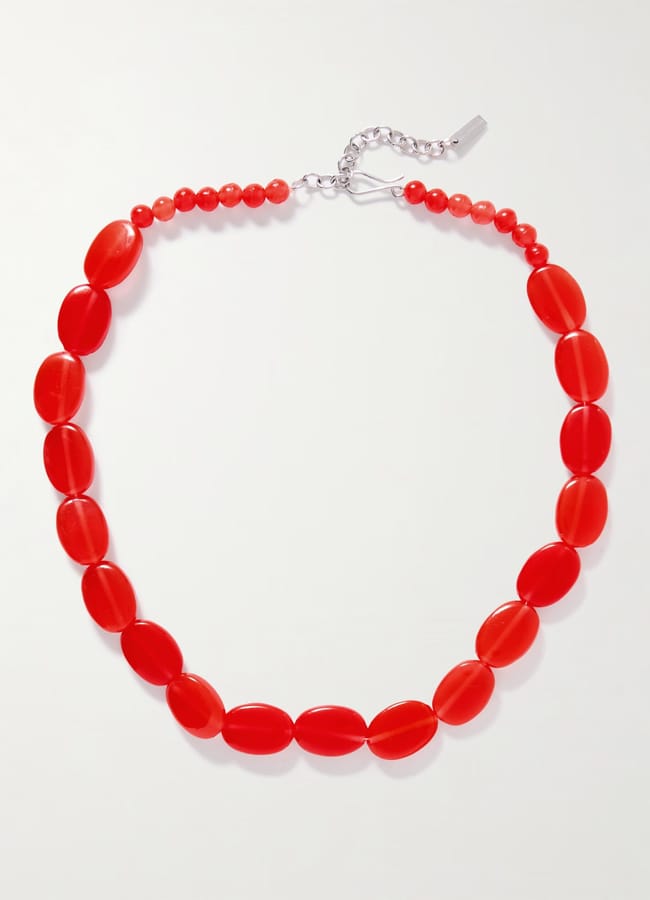 A necklace made of glossy oval and round red beads, arranged in a graduated pattern and connected by a silver chain with an adjustable clasp. This piece is a striking addition to any collection of red accessories. The background is plain white.