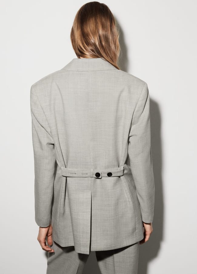 A person with long light brown hair, dressed in a light grey High Street blazer and matching trousers, stands with their back to the camera against a plain white background—a look fit for a Fashion Editor.