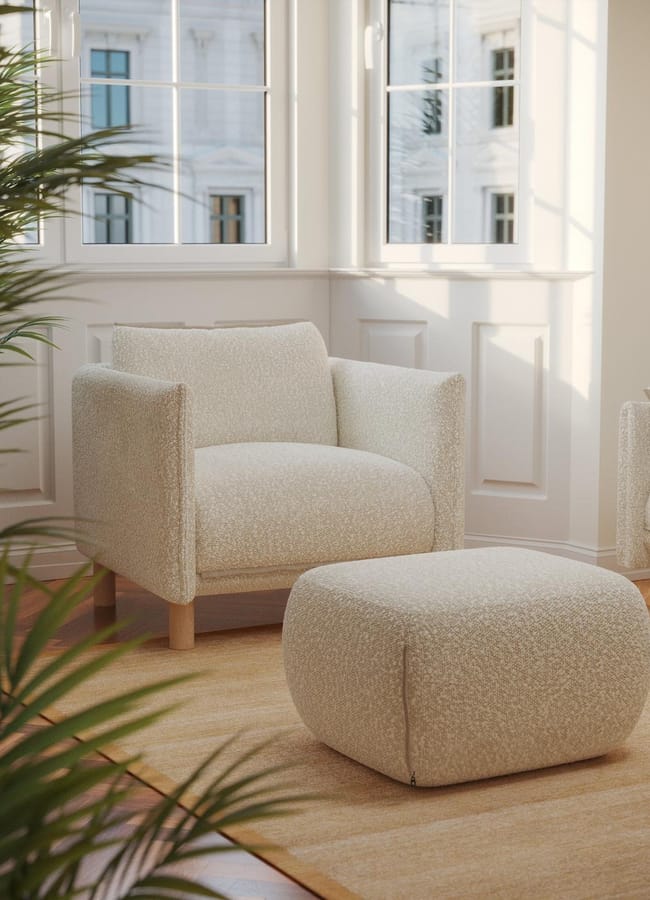 A cosy cream armchair and matching ottoman sit on a light rug in a bright room with large windows and white walls—a perfect example of the latest interiors trend in home décor, with sunlight streaming in and green leaves from a nearby plant in the foreground.