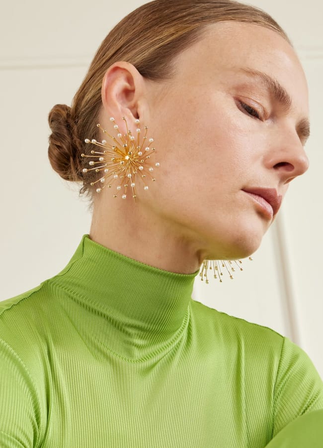 A woman with light skin and brown hair in a low bun wears a lime green ribbed polo neck—a nod to dopamine dressing—and large, gold, starburst-shaped earrings with pearl-like tips. She is looking downward with a neutral expression.