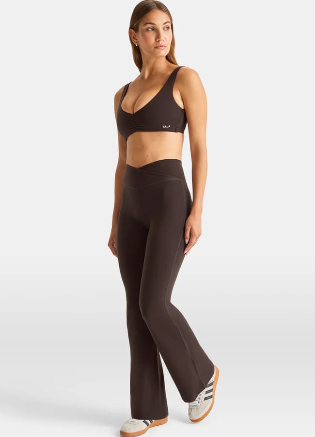 A woman stands wearing a dark brown athleisure sports bra and flared high-waisted leggings with white trainers, posing against a plain white background.