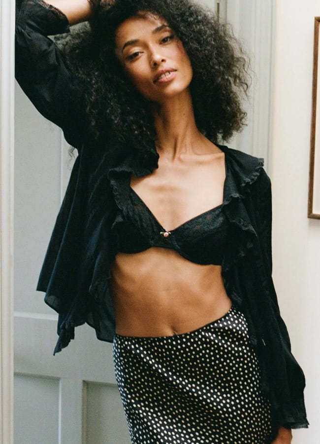 A woman with curly hair poses indoors in a black lingerie set, an open blouse, and a black skirt with white polka dots. She leans against a door frame with one arm raised, gazing confidently at the camera.