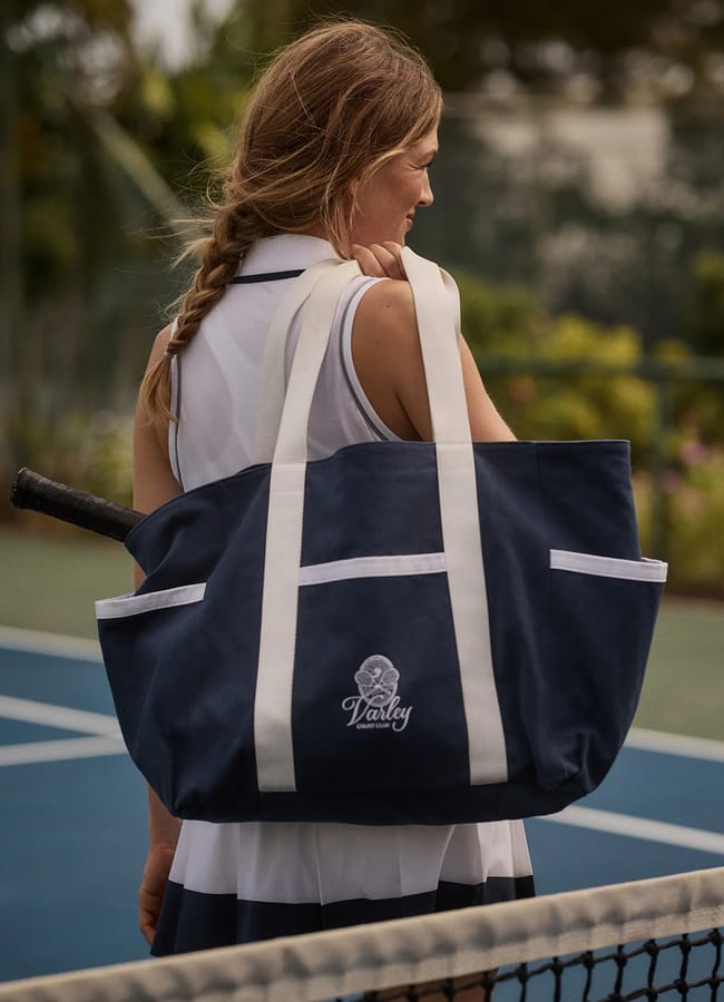 A woman in a white and navy athleisure tennis outfit stands on the court, carrying a large navy tote bag with white straps and a racquet handle visible. The bag features Varley and a logo printed on the front.