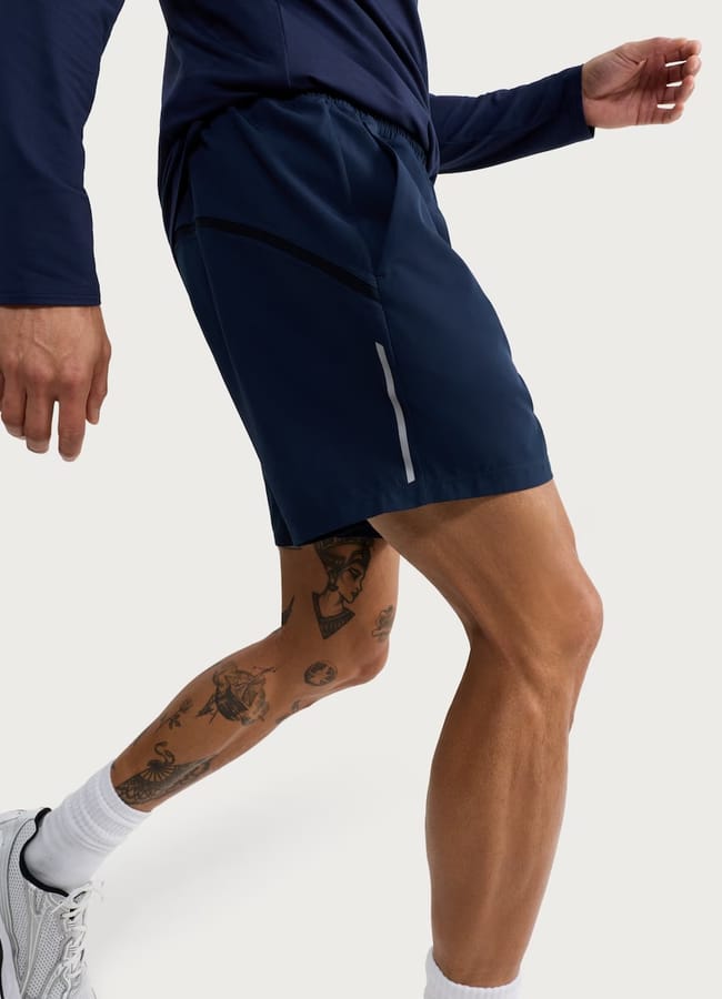 A person wearing men's gym clothes: a long-sleeved navy shirt, navy sports shorts with a reflective strip, white socks, and white trainers. Their tattooed right leg is slightly bent as they walk.