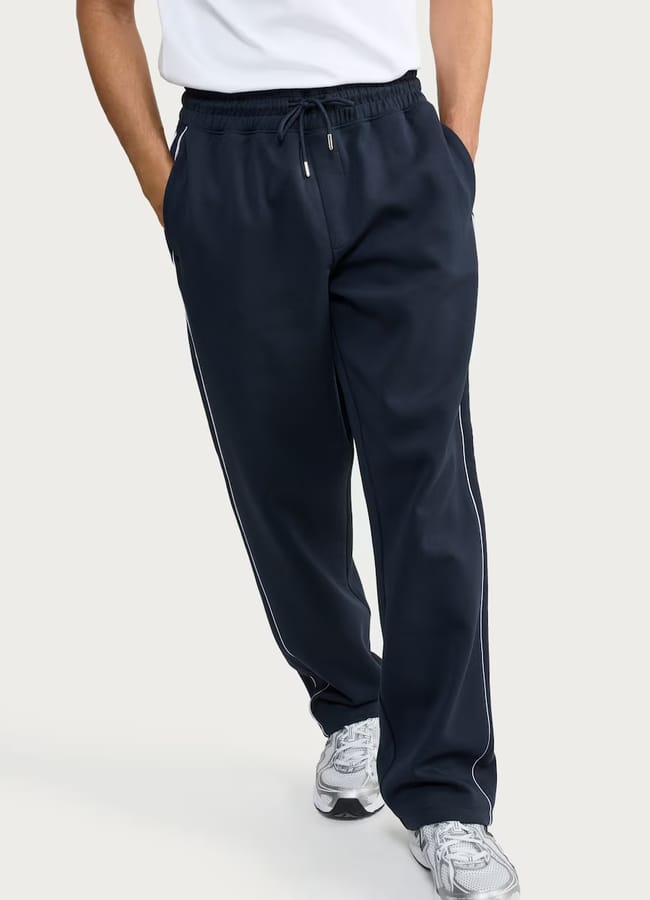 A person wearing gym clothes for men—navy blue tracksuit bottoms with white piping, a white shirt, and white trainers. With hands in pockets, he stands against a plain light background.