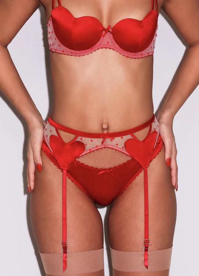 A person wearing a red lingerie set with heart-shaped details, matching suspender belt, and sheer tights stands confidently against a plain white background.