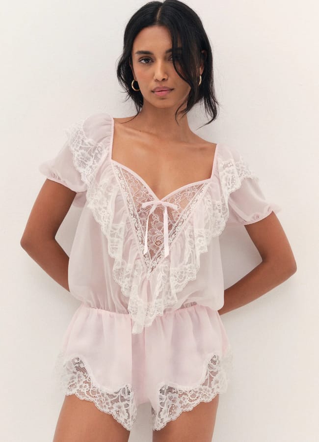 A woman poses against a white wall wearing a sheer, light pink playsuit with white lace trim, short sleeves, and frill details, styled like a delicate lingerie set. She has long dark hair, gold hoop earrings, and a calm expression.