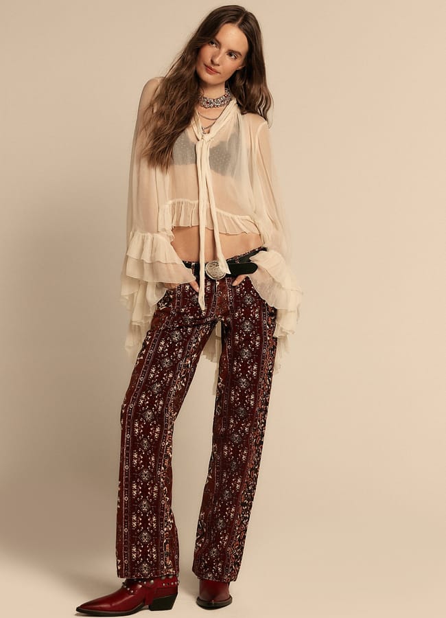 A woman embodies dopamine dressing in a sheer, ruffled cream blouse, black bralette, patterned maroon trousers, a belt, layered necklaces, and bold red heeled boots as she poses against a beige background.
