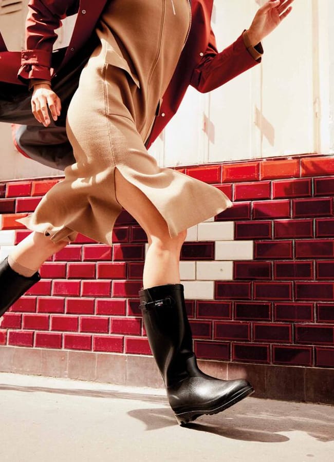 A person in a tan dress, red coat, and stylish black boots is captured mid-step walking past a wall with red and white tiles. Their fashionable footwear stands out, whilst their face is not visible in the image.