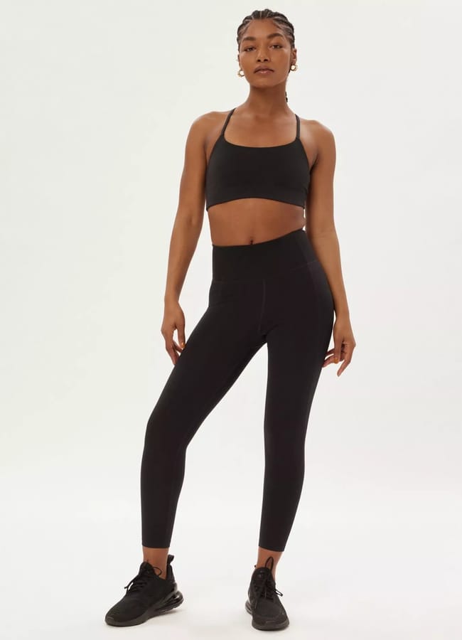 A woman wearing black athleisure—a sports bra, high-waisted leggings, and trainers—stands confidently against a plain white background.