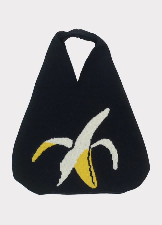 A black fabric tote bag with a simple, dopamine dressing-inspired graphic of a peeled banana in yellow and white on the front. The bag has a triangular shape and two handles at the top.