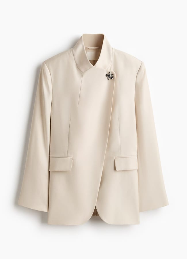 A cream-coloured, tailored High Street blazer for women with a high collar, asymmetric front, two front flap pockets, and a decorative brooch near the collar—worthy of any Fashion Editor. Displayed against a white background.