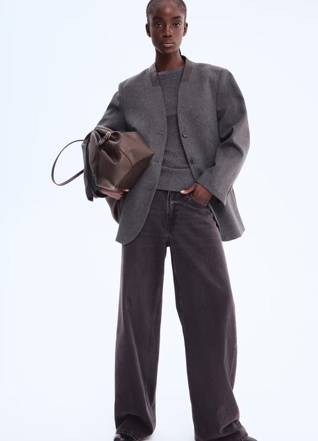 A person wearing a loose grey High Street blazer, matching jumper, and wide-leg dark jeans stands against a plain background, holding a large brown handbag and channelling confidence like a Fashion Editor.