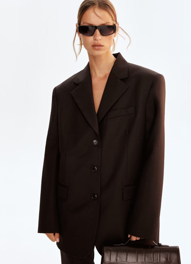 A woman styled like a Fashion Editor wears an oversized black High Street blazer, black sunglasses, and holds a dark textured handbag against a plain light background. Her hair is tied back with loose strands framing her face.