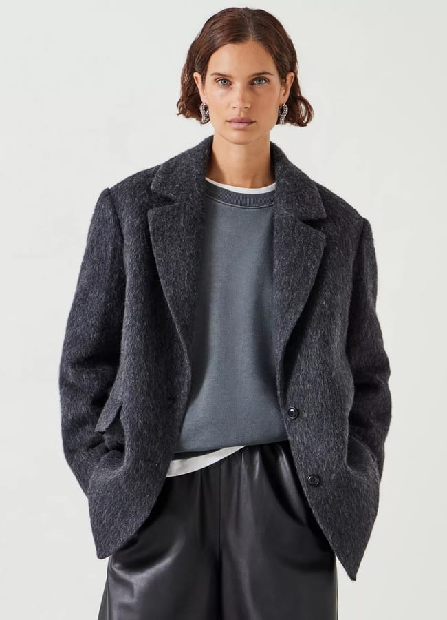 A woman with short brown hair, styled like a true fashion editor, wears a textured dark grey high street blazer over a grey sweatshirt and black leather trousers, standing against a plain light background.