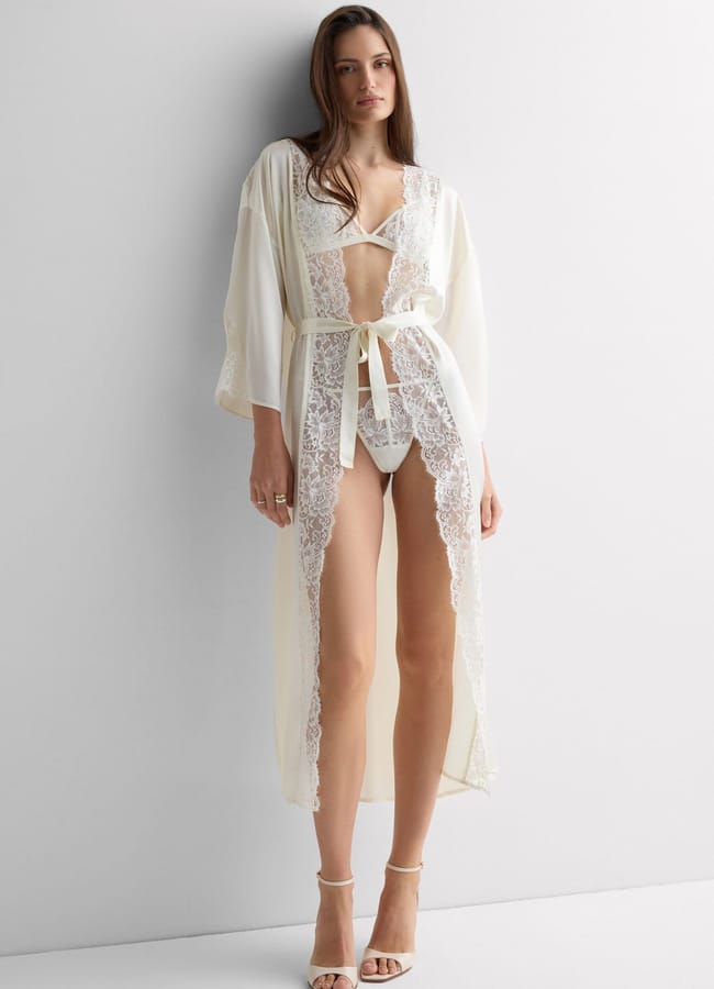 A woman stands against a white wall wearing a sheer, long, cream-coloured lace robe over a matching lingerie set and high-heeled sandals. She has long hair and a relaxed, confident pose.