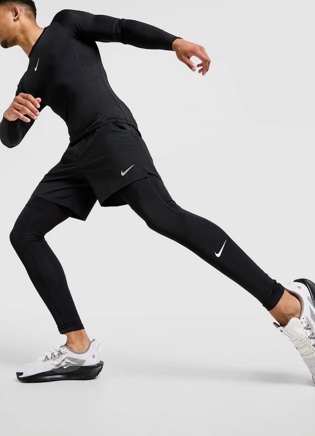 A person wearing black Nike gym clothes for men and white trainers is captured mid-stride against a plain light background, appearing to be in motion or running.