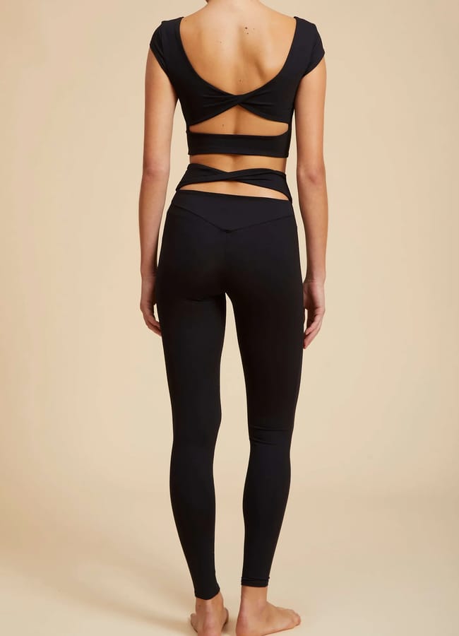 A woman stands barefoot facing away, dressed in a fitted athleisure black crop top with cut-outs across the back and matching high-waisted leggings, set against a plain beige background.