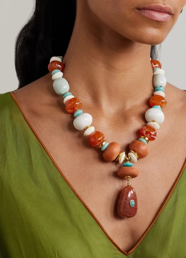 A woman in a green V-neck top embraces dopamine dressing, showcasing a chunky beaded necklace with large red, white, turquoise, and gold beads, finished with a teardrop-shaped red pendant at the centre.