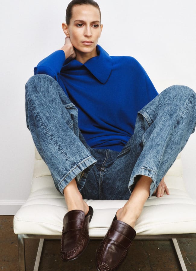 A person with slicked-back hair sits on a white chair, sporting dopamine dressing in a bright blue jumper, loose-fitting blue jeans, and brown leather loafers, with one hand resting on their neck and legs relaxed and apart.