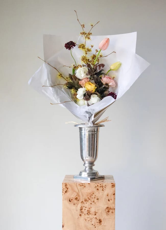 A bouquet of assorted Valentine’s Day flowers wrapped in white paper is arranged in a silver vase, placed on a wooden plinth against a plain light grey background.