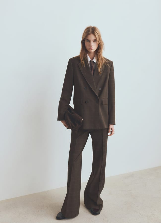 A woman stands against a light wall in a dark brown High Street blazer suit with wide-leg trousers, a white shirt, and brown tie. She holds a dark clutch, her long hair loose—a look worthy of any Fashion Editor.