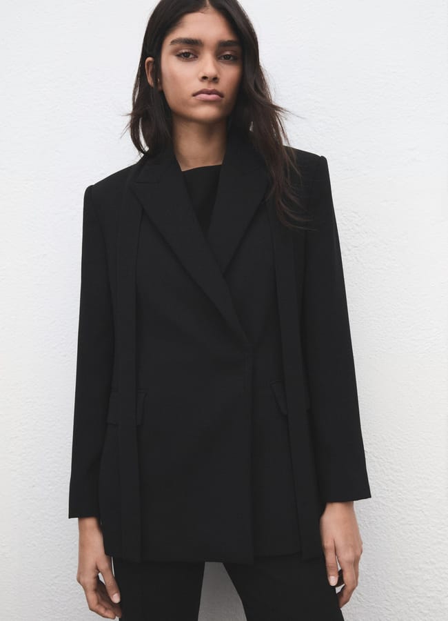 A woman with long dark hair, styled like a fashion editor, wears one of the best high street blazers—a black suit jacket over a black top—standing against a white textured wall, looking directly at the camera with a neutral expression.