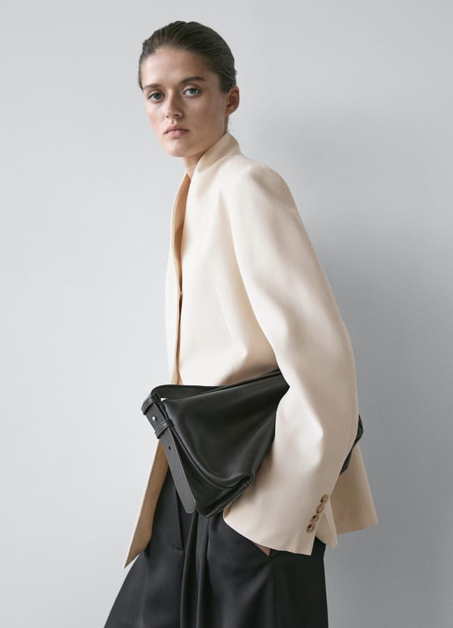 A person wearing a cream-coloured High Street blazer and black trousers poses against a plain light grey background, holding a large black clutch bag under one arm—an ensemble worthy of any Fashion Editor.