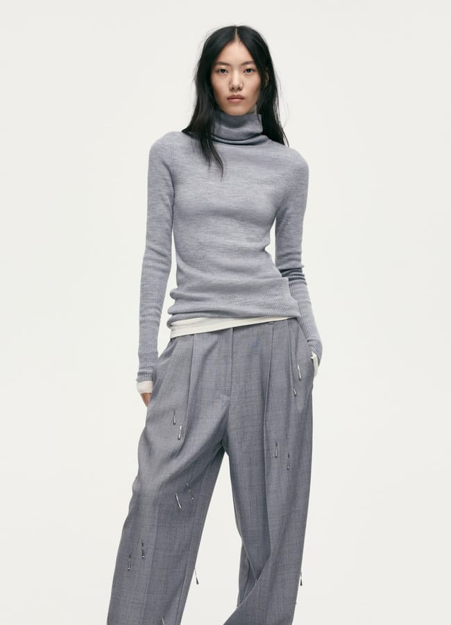 A woman with long black hair wears a light grey polo neck jumper and loose grey trousers with pleats and subtle pin details, embodying the minimalist style perfect for a No Spend Challenge, standing against a plain white background.