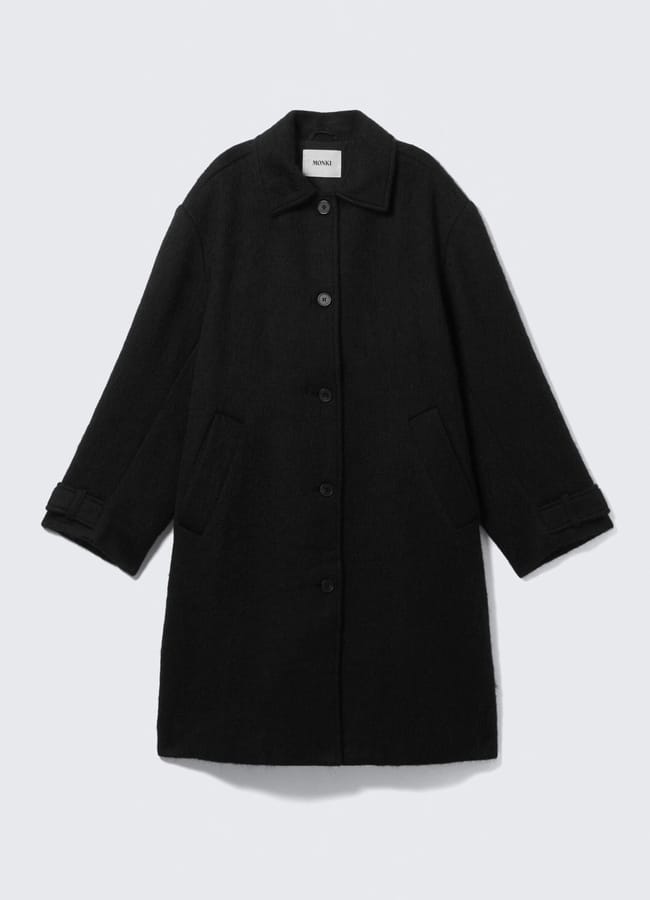 A long black wool coat with a collar, button-through front, side pockets, and adjustable straps on the cuffs—an elegant women’s workwear staple—displayed flat against a white background.