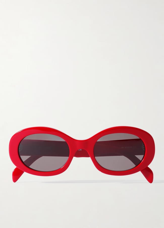 Bright red oval sunglasses with dark tinted lenses and thick frames, displayed against a plain light background. A bold choice for lovers of red accessories, the sunglasses are positioned with the arms slightly splayed.