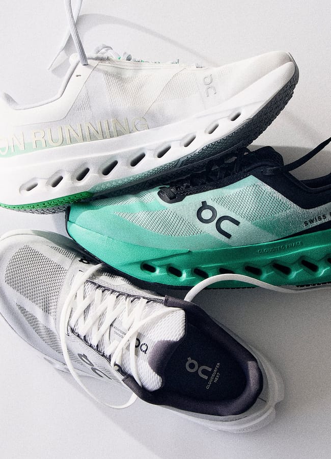Three pairs of On Running trainers in white, teal, and black are stacked on a white surface, showcasing the brand logo and unique sole design—perfect for completing your latest activewear look.
